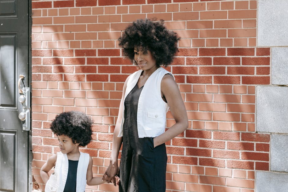 A mother and child in matching outfits stand in front of a brick wall, holding hands.