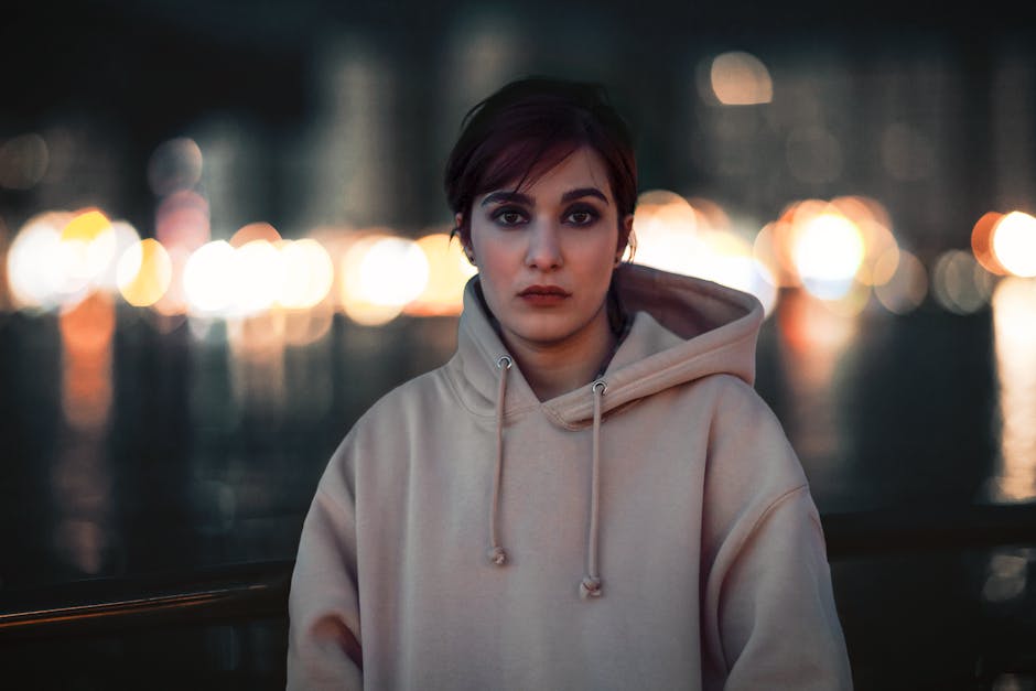 A woman in a beige hoodie standing outdoors with bokeh lights in the background at night.