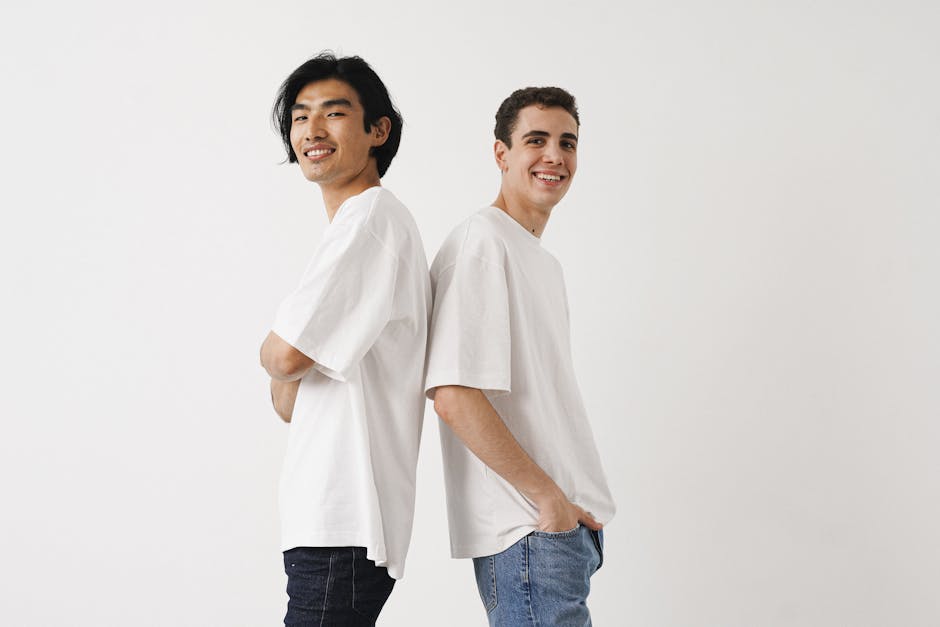 Two young men back to back in a studio, wearing white t-shirts and jeans, smiling confidently.