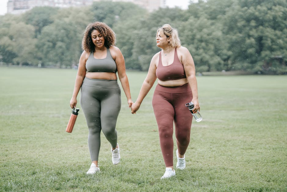 Two plus-size women in activewear holding hands while walking in the park, promoting fitness and friendship.